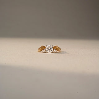 Front view of a round solitaire engagement ring in 14 KT Yellow Gold, featuring a round lab-grown diamond accented by sculpted leaf-inspired motifs and round side diamonds, displayed against a beige backdrop.