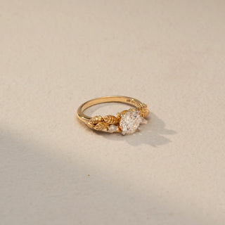 Side view of a round solitaire engagement ring in 14 KT Yellow Gold, featuring a round lab-grown diamond accented by sculpted leaf-inspired motifs and round side diamonds, displayed against a beige backdrop.
