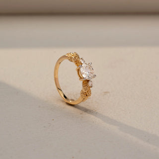 Angled view of a round solitaire engagement ring in 14 KT Yellow Gold, featuring a round lab-grown diamond accented by sculpted leaf-inspired motifs and round side diamonds, displayed against a beige backdrop.