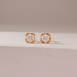 Front view of diamond stud earrings in 14KT yellow gold, featuring round brilliant diamonds set in floral-inspired four-petal settings, displayed on a beige surface highlighting classic sparkle and elegance.
