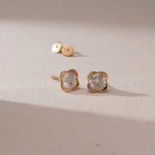 Angled view of diamond stud earrings in 14KT yellow gold, featuring round brilliant diamonds in floral-inspired four-petal settings, with butterfly backs on a beige surface highlighting soft sparkle.
