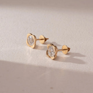 Side view of diamond stud earrings in 14KT yellow gold, featuring round brilliant diamonds in floral-inspired four-petal settings with screw backs, displayed on a beige surface highlighting warm glow and sparkle.
