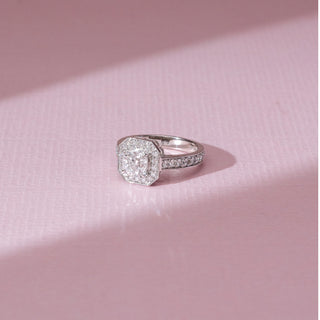 Angled view of halo diamond ring in white gold, featuring a square-cut lab-grown diamond encircled by a double halo with a diamond-studded band. Set on a soft pink surface, it reflects refined sparkle.
