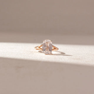 Front view of emerald-cut diamond solitaire ring in yellow gold with a split-style band, set on a soft neutral surface, highlighting elongated facets and clean, radiant brilliance.
