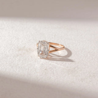 Side view of emerald-cut diamond solitaire ring in yellow gold with a split band, set on a soft neutral background, highlighting elongated facets and refined, radiant brilliance.
