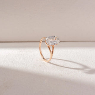 Angled view of emerald-cut diamond solitaire ring in yellow gold with a split band, set on a soft neutral background, highlighting elevated setting and clean, radiant brilliance.

