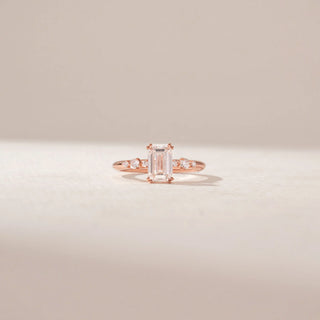 Front view of an emerald-cut diamond ring in 14KT Rose Gold, featuring a split band with round diamond accents against a beige backdrop.
