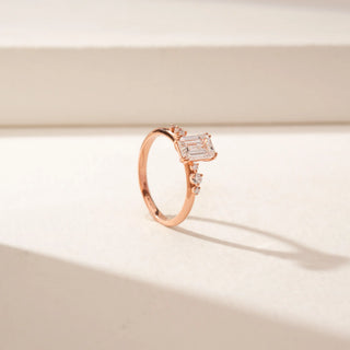 Angled view of an emerald-cut diamond ring in 14KT Rose Gold, featuring a split band with round diamond accents against a beige backdrop.
