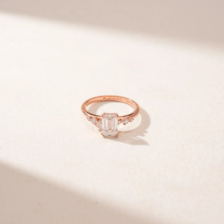 Top view of an emerald-cut diamond ring in 14KT Rose Gold, featuring a split band with round diamond accents against a beige backdrop.