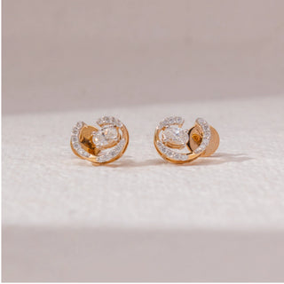 Front view of spiral diamond stud earrings in yellow gold, featuring centred round lab-grown diamonds surrounded by curved pavé-set arcs. Placed on a textured beige surface, the design reflects modern elegance.
