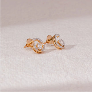 Angled view of spiral diamond stud earrings in yellow gold, featuring centred round lab-grown diamonds encircled by curved pavé-set arcs. Placed on a textured beige surface, the design casts a soft shadow.
