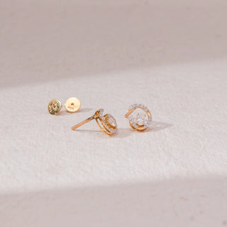 Top view of spiral diamond stud earrings in yellow gold, featuring centred round lab-grown diamonds with curved pavé-set arcs. Placed on a textured beige surface, the design casts a soft shadow.
