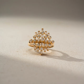Front view of a geometric diamond statement ring in 14 KT Yellow Gold, featuring round and baguette-cut lab-grown diamonds arranged in an intricate lattice-inspired design, displayed against a beige backdrop.