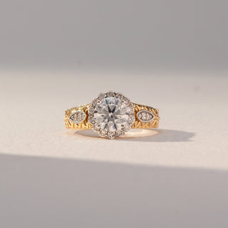 Front view of Vintage Halo Diamond Ring in 14KT Yellow Gold, featuring a round lab grown diamond with halo and engraved band details. Set on a beige backdrop, the design highlights intricate elegance and brilliance.
