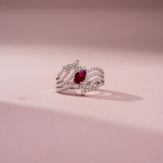 Front view of a gemstone statement ring in White Gold, featuring an oval-cut ruby-red center stone surrounded by flowing pavé-set diamond ribbons in an intricate openwork design, displayed against a soft pink backdrop.