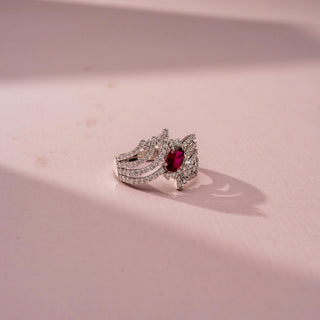 Side view of a gemstone statement ring in White Gold, featuring an oval-cut ruby-red center stone surrounded by flowing pavé-set diamond ribbons in an intricate openwork design, displayed against a soft pink backdrop.