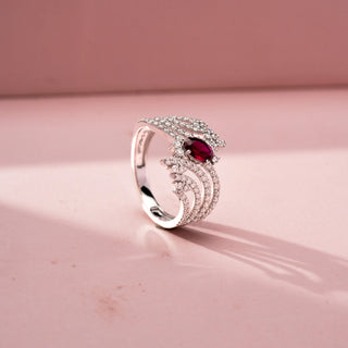Angled view of a gemstone statement ring in White Gold, featuring an oval-cut ruby-red center stone surrounded by flowing pavé-set diamond ribbons in an intricate openwork design, displayed against a soft pink backdrop.