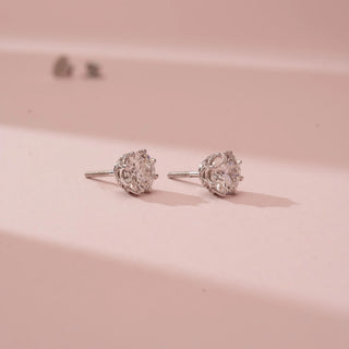 Angled view of Reia’s 14 KT White Gold Classic Round Brilliant Diamond Studs. It highlights the round brilliant-cut diamonds in architectural multi-prong settings against a soft pink backdrop.