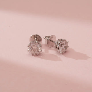 Top-down view of Reia’s 14 KT White Gold Classic Round Brilliant Diamond Studs. The layout highlights the round-cut diamonds and architectural multi-prong settings against a soft pink backdrop.