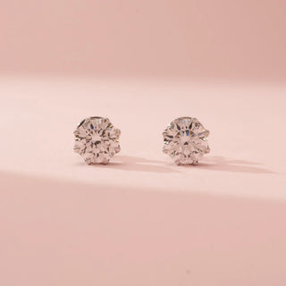 Front view of Reia’s 14 KT White Gold Classic Round Brilliant Diamond Studs. The layout highlights the round brilliant-cut diamonds in refined multi-prong settings against a soft pink backdrop.