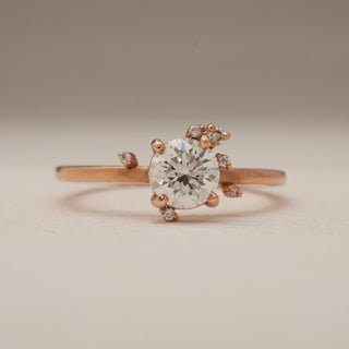 Front close-up of Whimsical Floral Diamond Engagement Ring crafted in 14 KT rose gold, showcasing a round solitaire of 0.69 carats accented with tiny floral side diamonds, photographed on a flat beige surface.