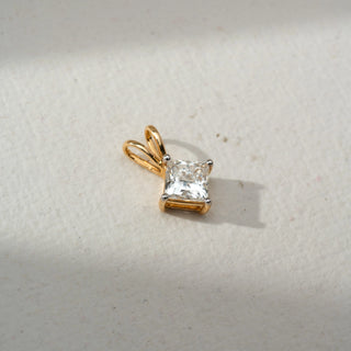 Front view of a yellow gold pendant featuring a princess-cut solitaire diamond in a four-prong setting, displayed on a soft neutral surface with a polished double-loop bail.