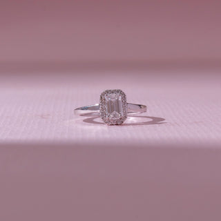 Front view of an emerald-cut halo diamond ring in white gold, featuring a centred emerald-cut diamond framed by pavé stones. Captured on a soft pink backdrop, highlighting vintage elegance.
