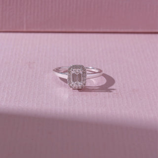 Top view of an emerald-cut halo diamond ring in white gold, featuring a centred emerald-cut diamond framed by pavé stones. Captured on a soft pink backdrop, highlighting timeless sparkle.
