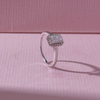 Angled view of an emerald-cut halo diamond ring in white gold, featuring a centred emerald-cut diamond framed by pavé stones. Captured on a soft pink backdrop, highlighting elegant brilliance.
