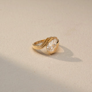 Side view of a pear solitaire ring in 14 KT Yellow Gold, featuring a pear-shaped lab-grown diamond embraced by flowing sculpted gold curves in an elegant bypass design, displayed against a beige backdrop.