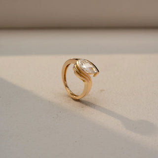 Side front view of a pear solitaire ring in 14 KT Yellow Gold, featuring a pear-shaped lab-grown diamond embraced by flowing sculpted gold curves in an elegant bypass design, displayed against a beige backdrop.