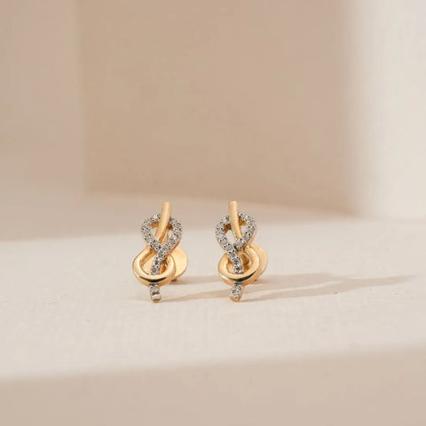 Front view of infinity loop diamond earrings in yellow gold featuring intertwined curves with round lab grown diamond accents and a delicate drop detail, placed on a soft neutral surface.