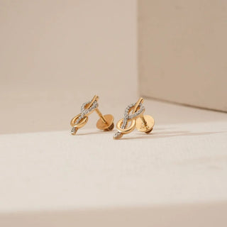 Side view of infinity loop yellow gold diamond earrings featuring intertwined curves with round lab grown diamond accents and a small drop detail, displayed on a soft neutral studio surface.