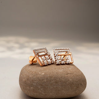 Angled view of geometric diamond stud earrings in 14 KT Rose Gold, featuring pavé-set rectangular frames with floral diamond clusters, displayed on a textured stone surface.

