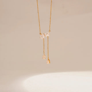 Front view of a 14KT yellow gold necklace featuring emerald and pear-cut diamonds in a drop design, set against a beige background.