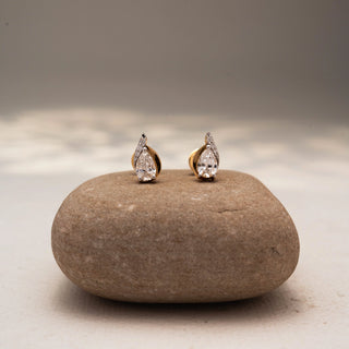 Front view of pear-shaped diamond stud earrings in 14 KT Yellow Gold, featuring bezel-set pear diamonds with pavé accents, styled on a textured stone surface.