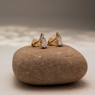 Side view of pear-shaped diamond stud earrings in 14 KT Yellow Gold, featuring bezel-set pear diamonds with pavé accents, displayed on a textured stone surface.