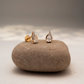 Angled view of pear-shaped diamond stud earrings in 14 KT Yellow Gold, featuring bezel-set pear diamonds with pavé accents, displayed on a textured stone surface.