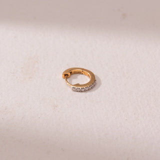 Top view of a gold nose ring, featuring a row of pavé-set diamonds along the front curve. Captured against a soft beige backdrop, the design reflects delicate sparkle and minimal elegance.

