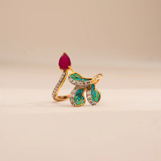 Angled front view of Reia’s 14 KT Yellow Gold Elegant Ruby Enamel Diamond Ring. It highlights the vibrant teal enamel, a pear-shaped ruby center stone, and shimmering round diamonds on a cream backdrop.