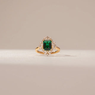 Close-up view of an emerald-cut green gemstone ring in 14KT Yellow Gold, featuring a diamond halo and marquise accents against a beige backdrop.
