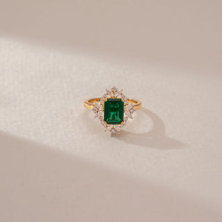 Front view of an emerald-cut green gemstone ring in 14KT Yellow Gold, featuring a diamond halo and marquise accents against a beige backdrop.