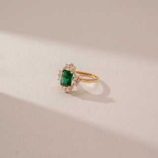 Side view of an emerald-cut green gemstone ring in 14KT Yellow Gold, featuring a diamond halo and marquise accents against a beige backdrop.