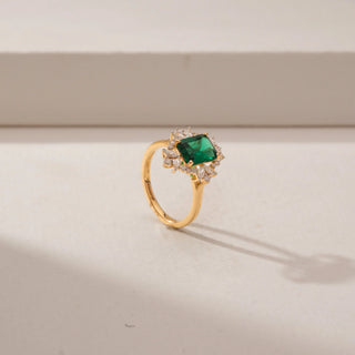 Angled view of an emerald-cut green gemstone ring in 14KT Yellow Gold, featuring a diamond halo and marquise accents against a beige backdrop.