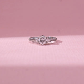 Front view of three-stone diamond ring in white gold, featuring a centred round lab-grown diamond flanked by two smaller stones and a pavé-set band. Set against a soft pink backdrop, the design reflects elegant brilliance.