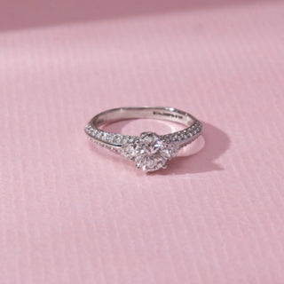 Top view of three-stone diamond ring in white gold, featuring a larger centre round lab-grown diamond flanked by two smaller stones on a pavé band. Placed on a soft pink surface, the design casts a gentle shadow.
