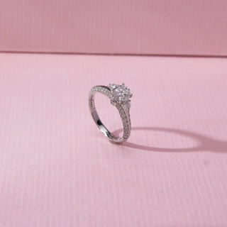 Side view of three-stone diamond ring in white gold, featuring a larger centre round lab-grown diamond with two smaller stones and a pavé band. Set on a soft pink surface, the design casts a delicate shadow.

