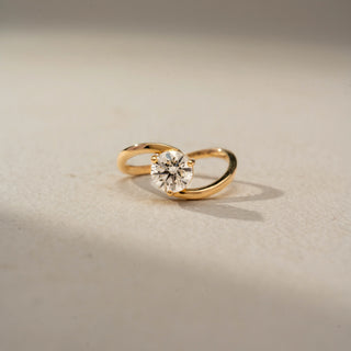 Front view of a round solitaire engagement ring in 14 KT Yellow Gold, featuring a round lab-grown diamond embraced by a sleek open bypass band, displayed against a beige backdrop.