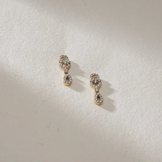 Pair of double drop diamond dangle earrings in 14KT Yellow Gold, placed on a textured surface, featuring interlinked teardrop motifs with EF-VVS round diamonds.