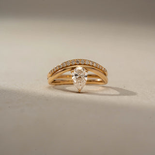 Front view of a pear solitaire ring in 14 KT Yellow Gold, featuring a bezel-set pear-shaped lab-grown diamond paired with a pavé-set curved band in a modern stacked design, displayed against a beige backdrop.